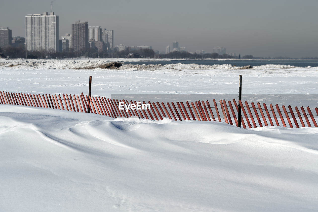 Snow covered land with snowdrifts and snow fence against sky