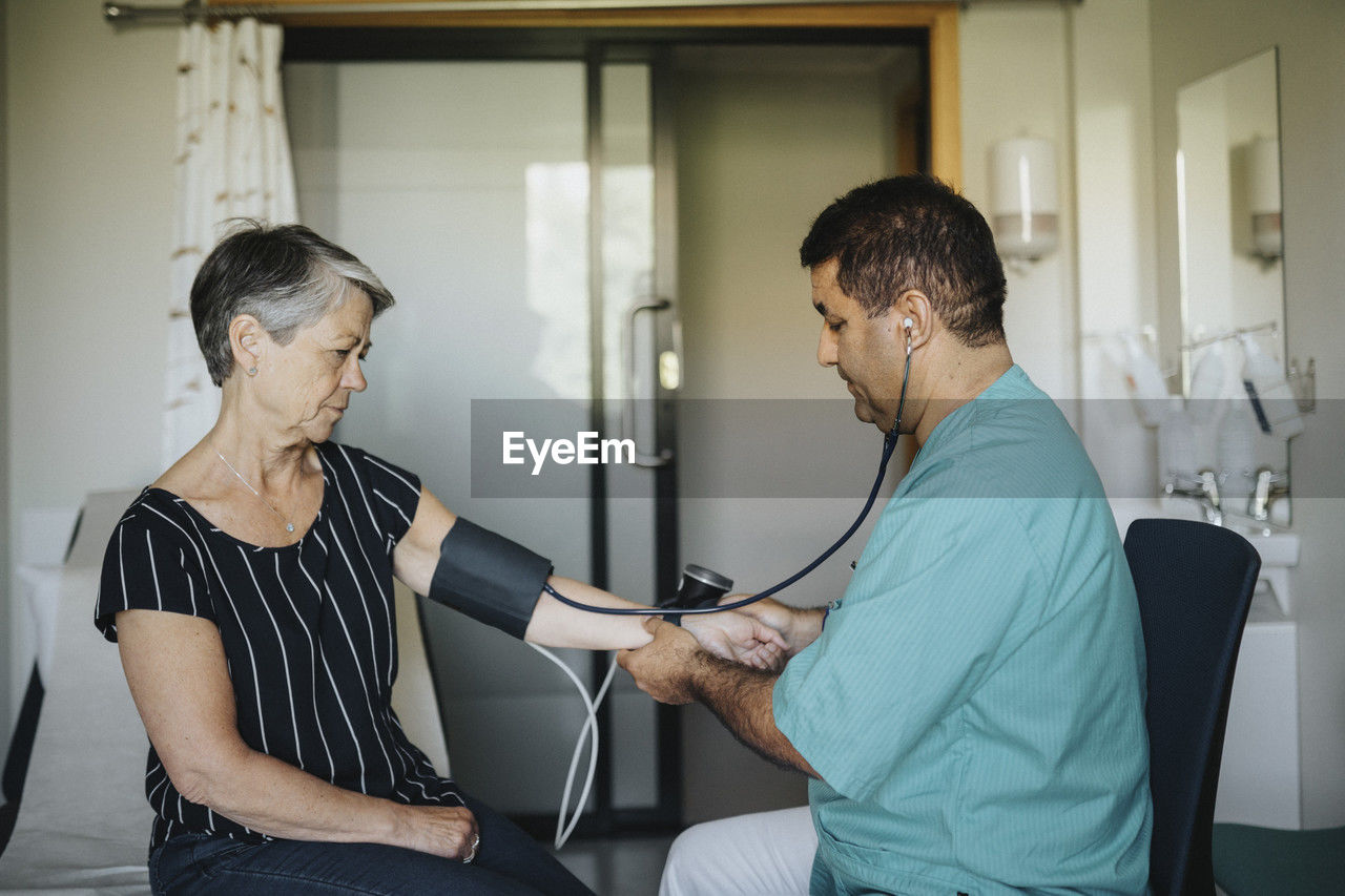 Male doctor checking blood pressure of senior woman while sitting in hospital