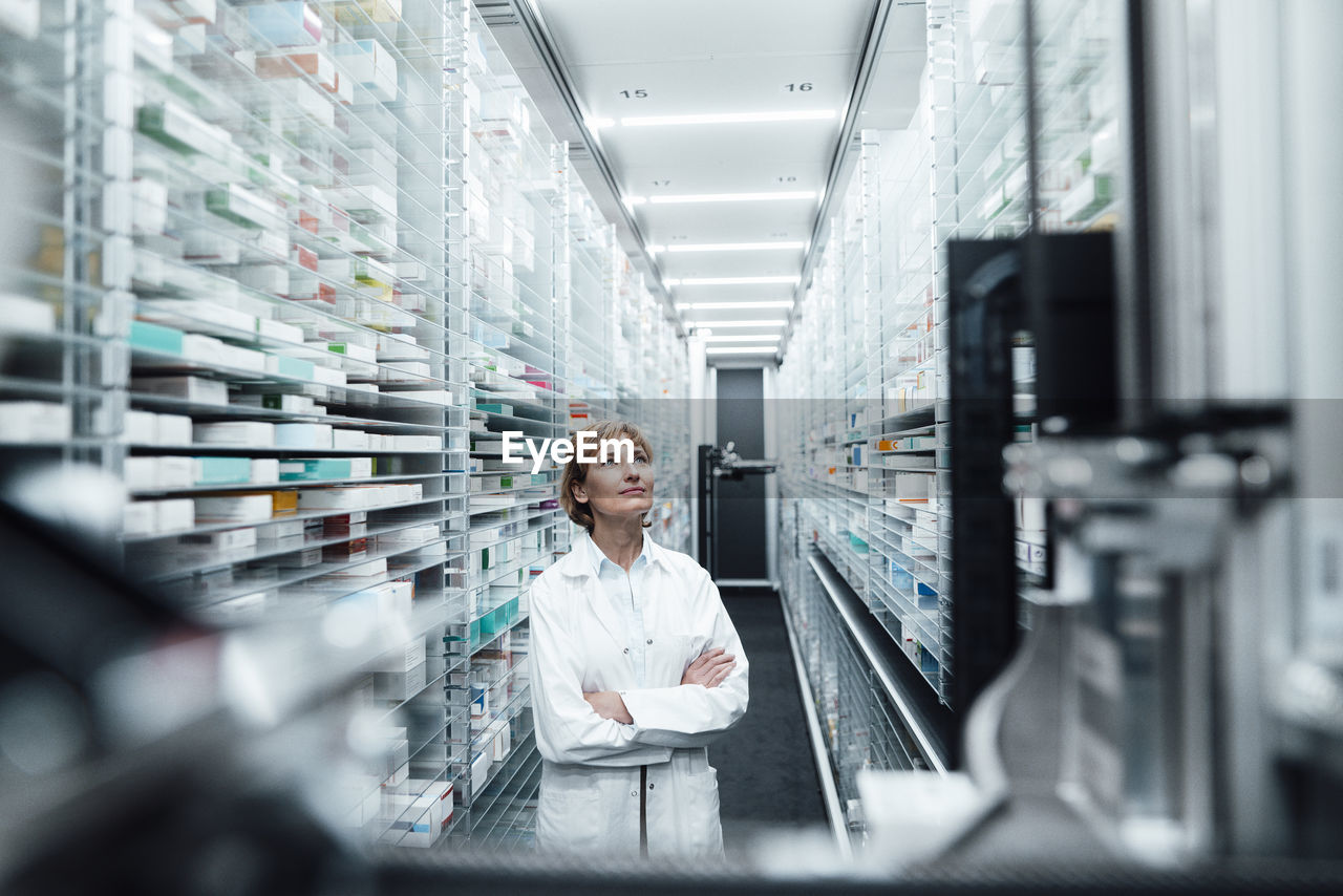 Female pharmacist with arms crossed looking at medicine in pharmacy store