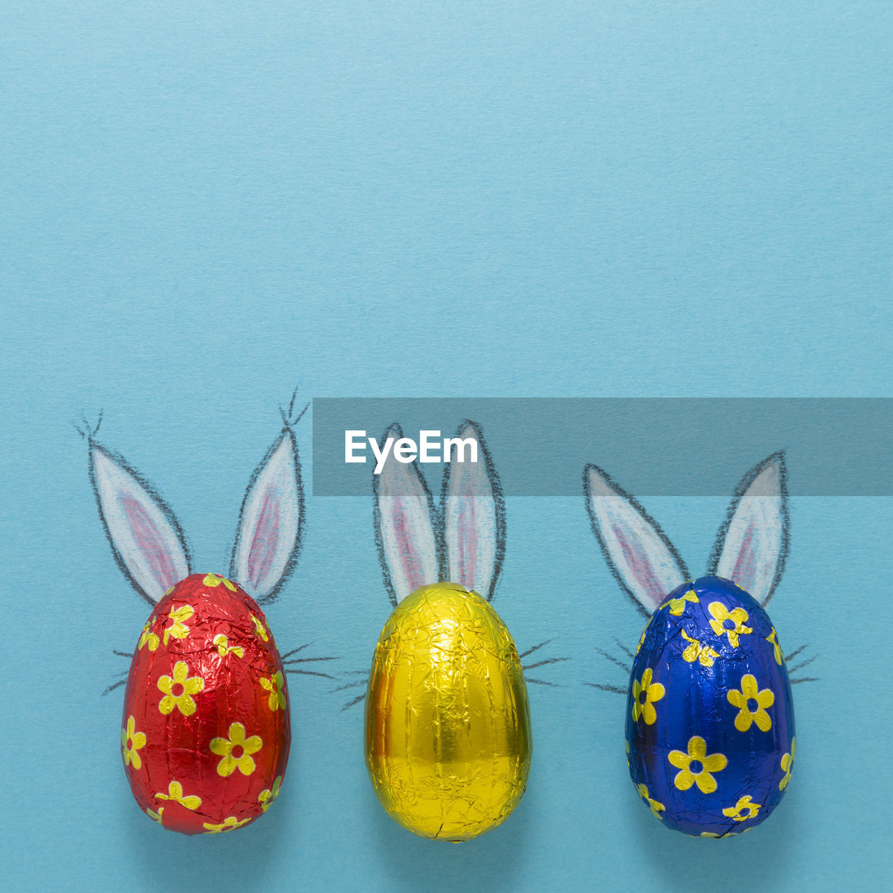 Three chocolate eggs in colorful aluminium foil with painted easter bunny ears on blue background