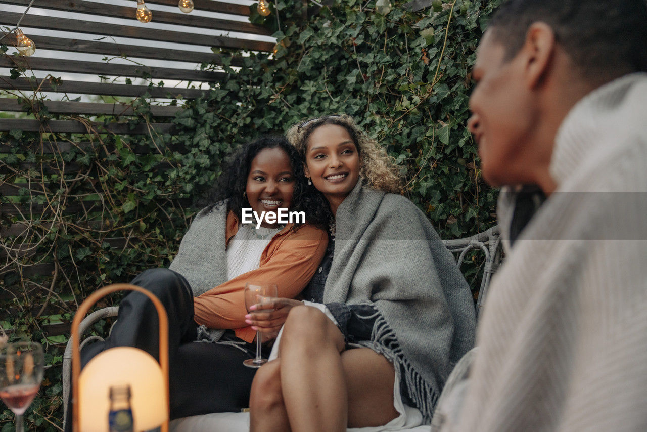 Smiling female friends wrapped in blanket talking with male friend in back yard