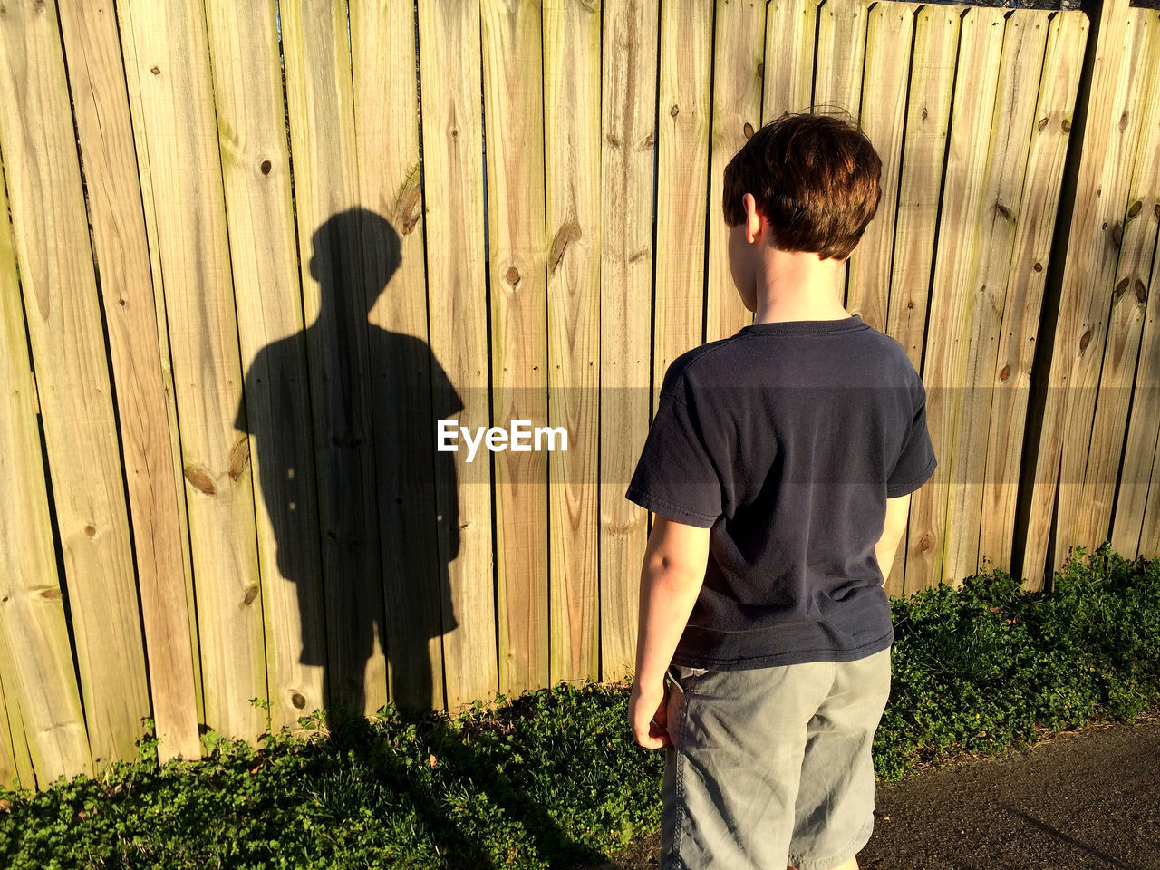 Rear view of boy standing by wooden fence on sunny day