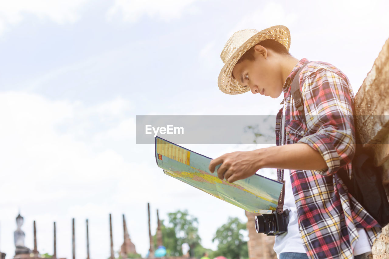 Low angle view of young man looking at map while standing against sky