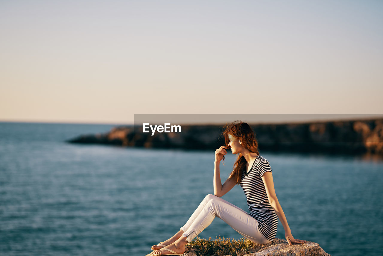 WOMAN SITTING BY SEA AGAINST SKY