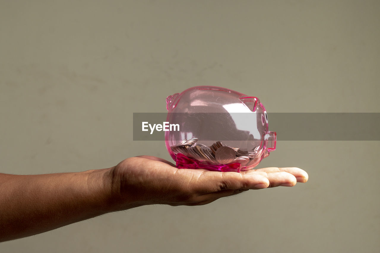 Cropped image of hand holding piggy bank against gray background