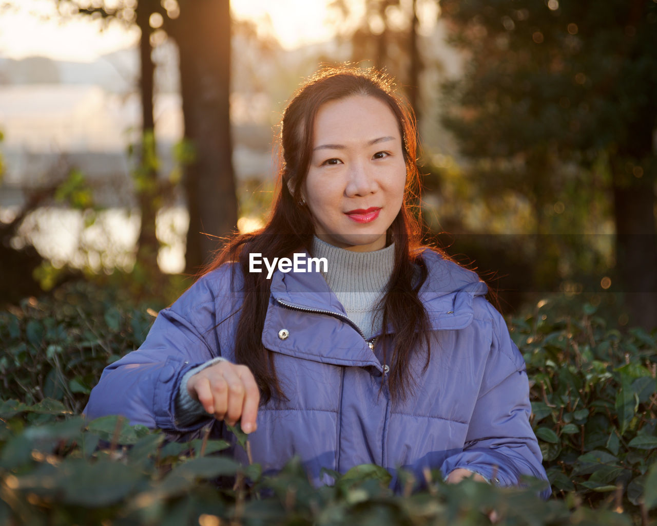 Portrait of smiling mid adult woman in warm clothing standing amidst plants in park during sunset