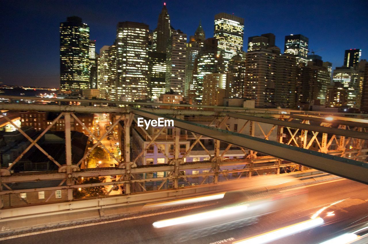 LIGHT TRAILS ON BRIDGE AGAINST BUILDINGS AT NIGHT