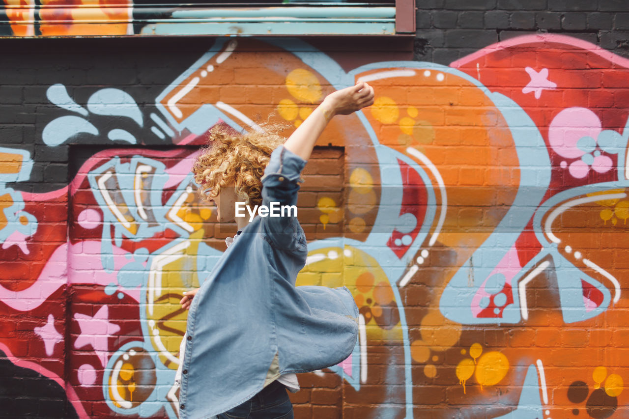 Young woman standing with hand raised against graffiti wall