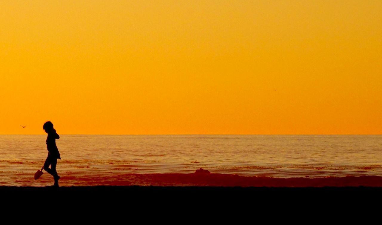 Silhouette child walking at beach against orange sky during sunset