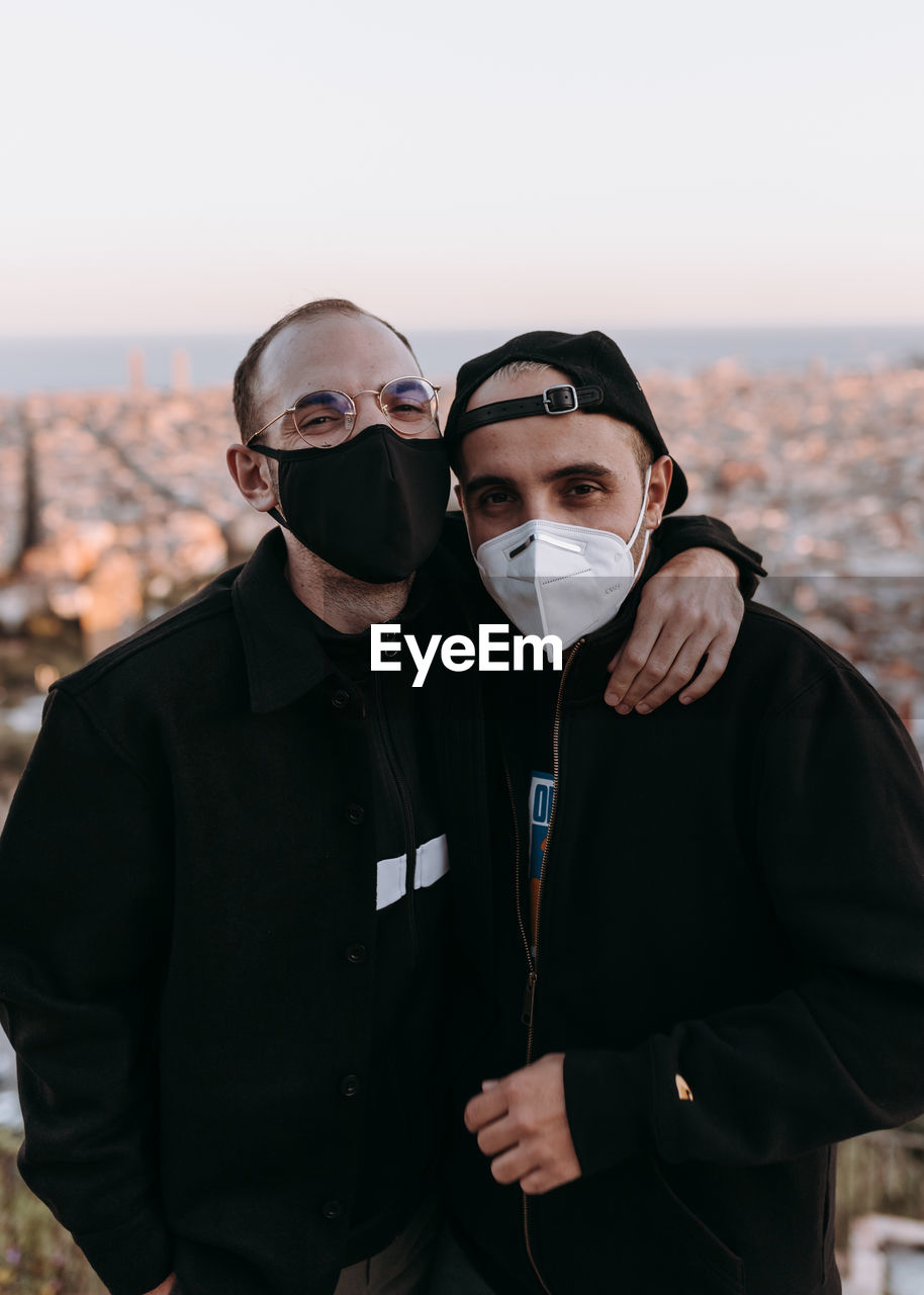 Portrait of two young men embracing and wearing face masks standing against cityscape