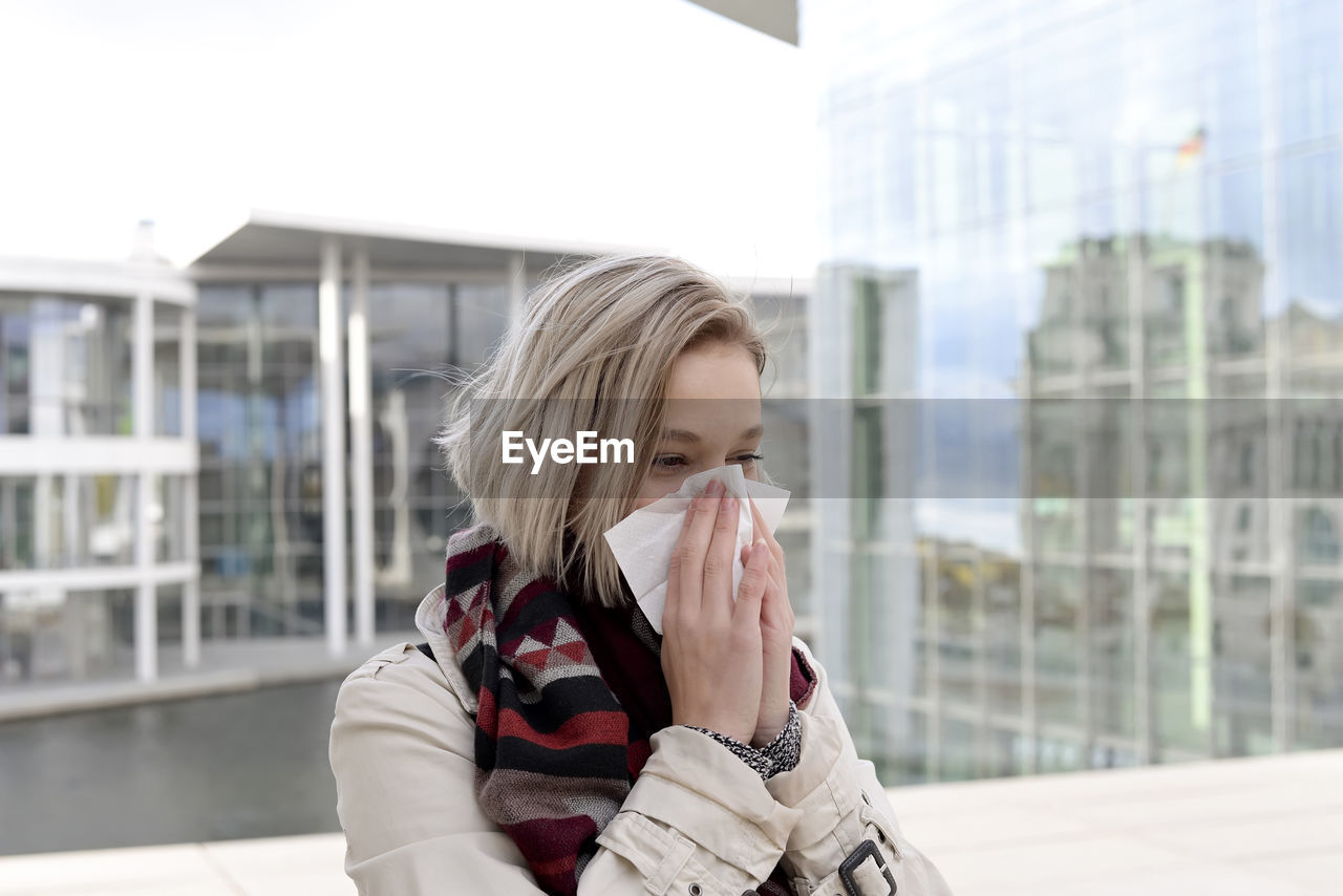 Young woman blowing nose while standing against building 