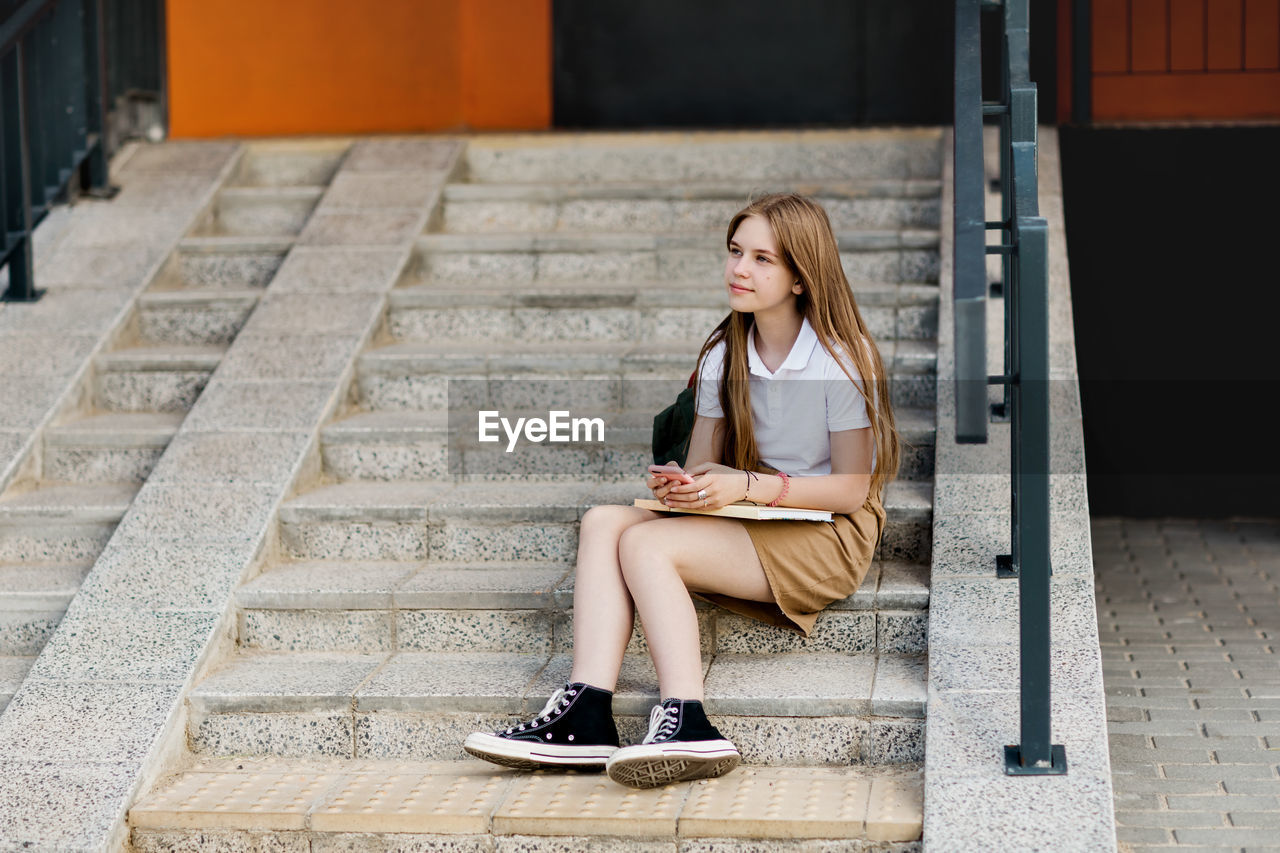 Cute girl sitting on the stairs outside the campus after college class and chatting with her friends