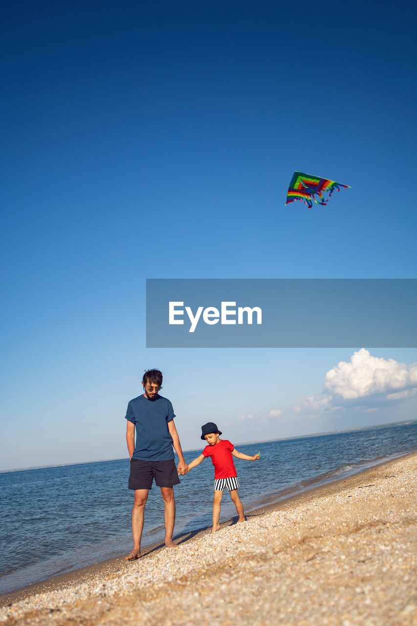 Father and son are standing on a sandy beach by the sea and launch a toy striped kite in the summer