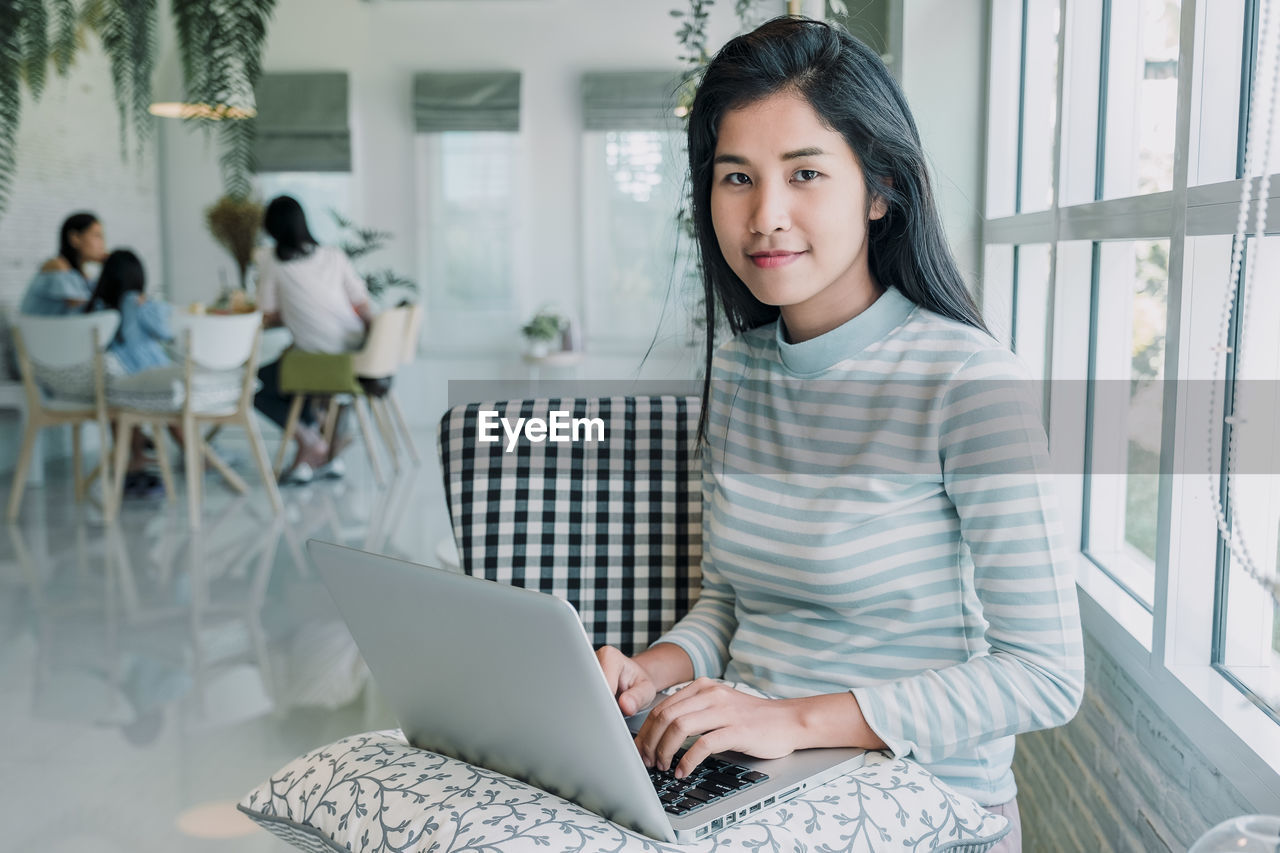 PORTRAIT OF SMILING YOUNG WOMAN USING PHONE WHILE SITTING IN LAPTOP