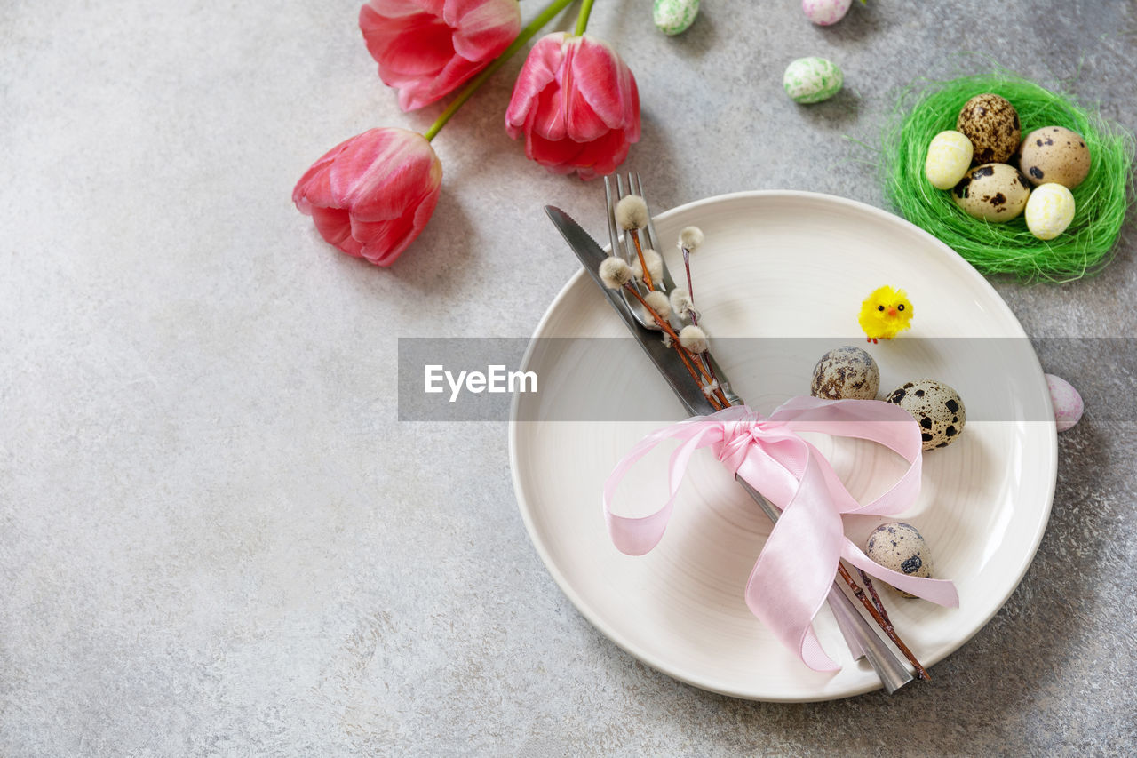 Festive easter table setting with painted eggs, spring flowers and cutlery on light grey tabletop. 