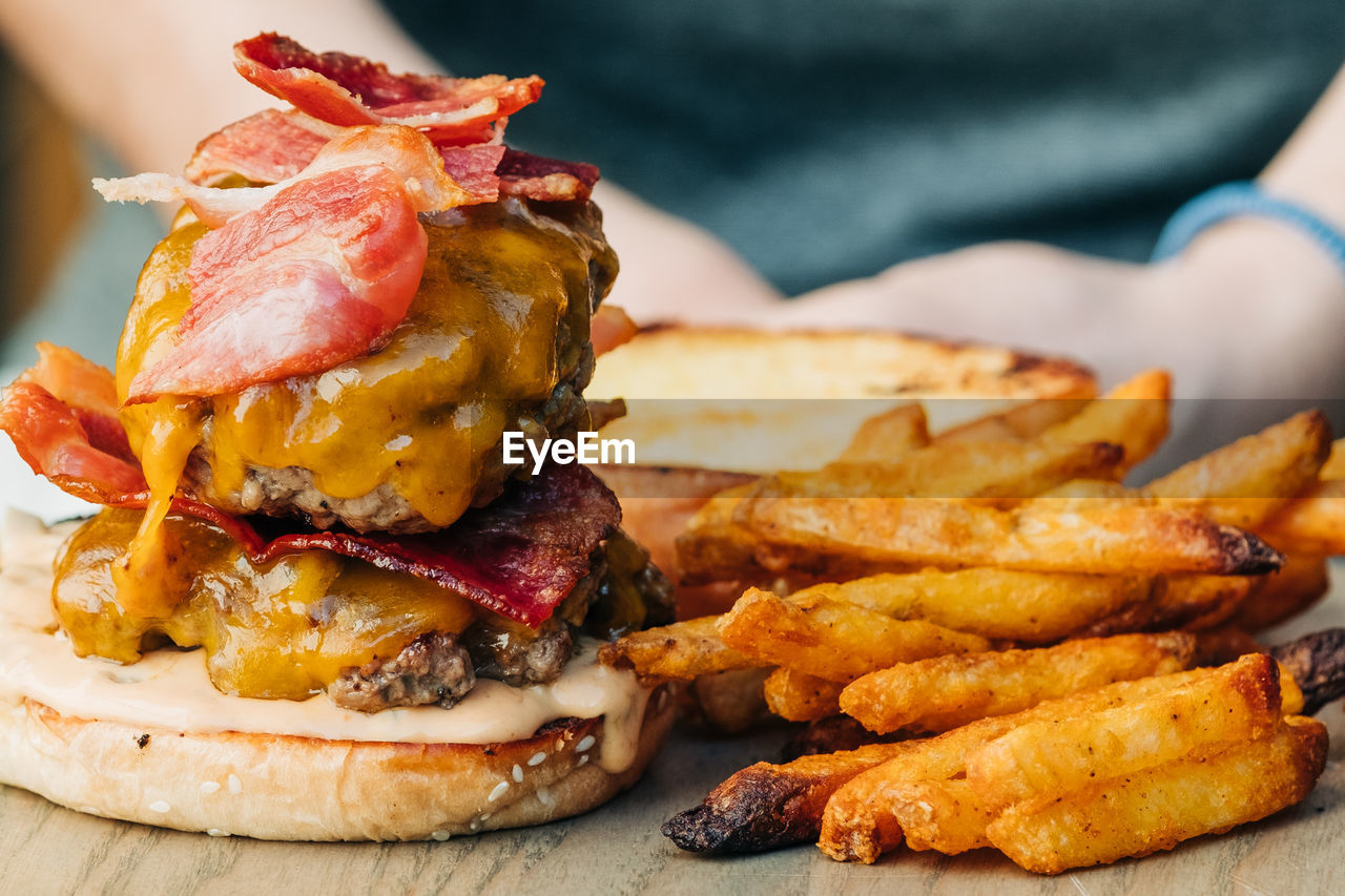 Close-up of double cheeseburger with bacon next to french fries on wooden board