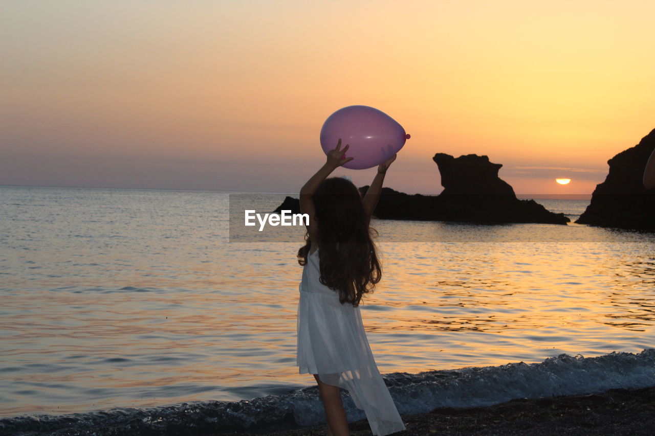 Girl holding balloon while standing by sea against sky during sunset