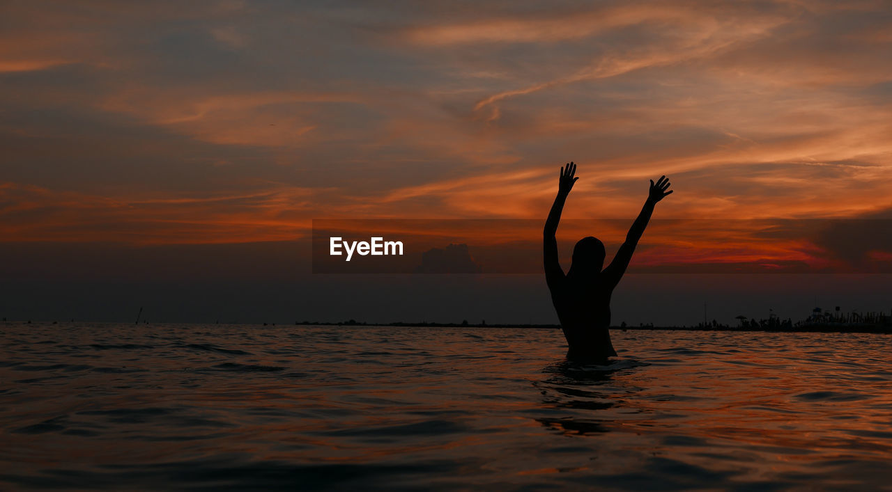 Silhouette woman with arms raised swimming in sea against sky during sunset