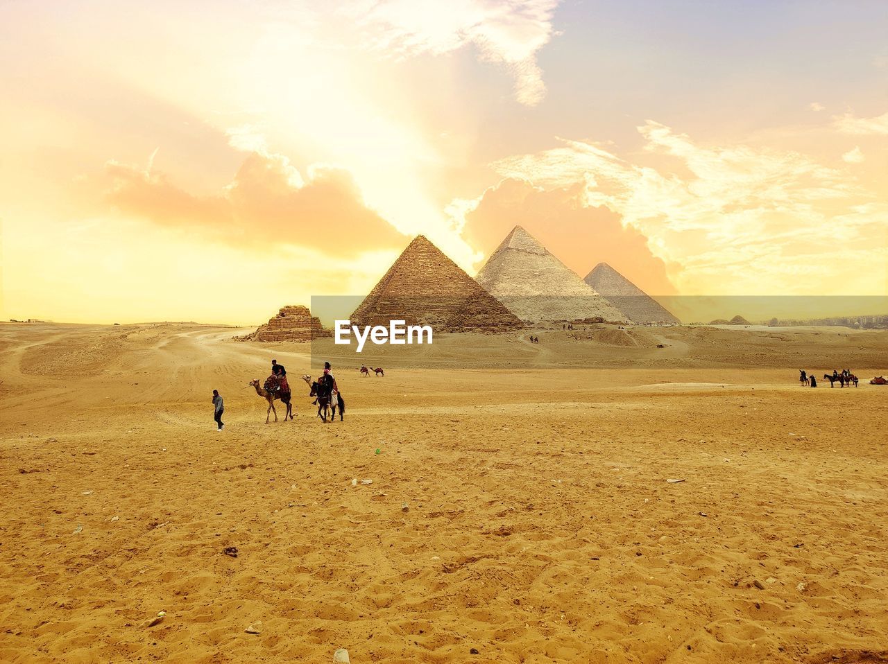 desert, sky, camel, travel destinations, land, domestic animals, pyramid, mammal, travel, landscape, nature, scenics - nature, sand, animal themes, animal, ancient, history, natural environment, environment, the past, tourism, architecture, cloud, sunset, beauty in nature, pet, ancient civilization, sunlight, climate, arid climate, group of people, group of animals, wadi, monument, plain, outdoors, men, horizon, city, tranquil scene, adult, working animal, plateau, built structure, day, tranquility, tourist, archaeology, sun, journey, walking, erg