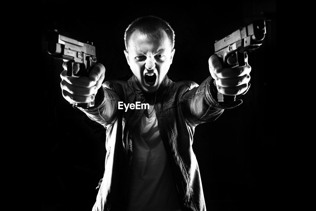 Angry man aiming with guns while standing against black background