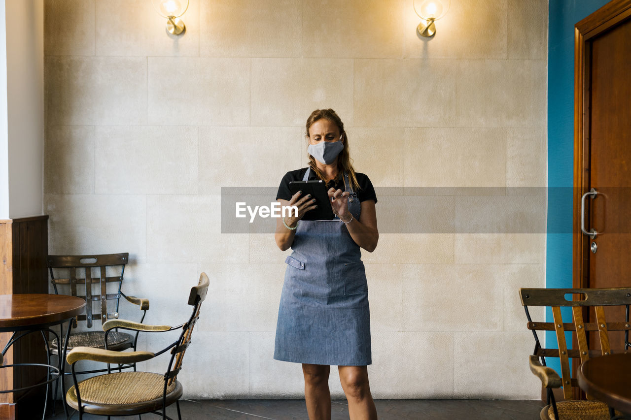 Female cafe owner wearing protective face mask while using digital tablet against wall