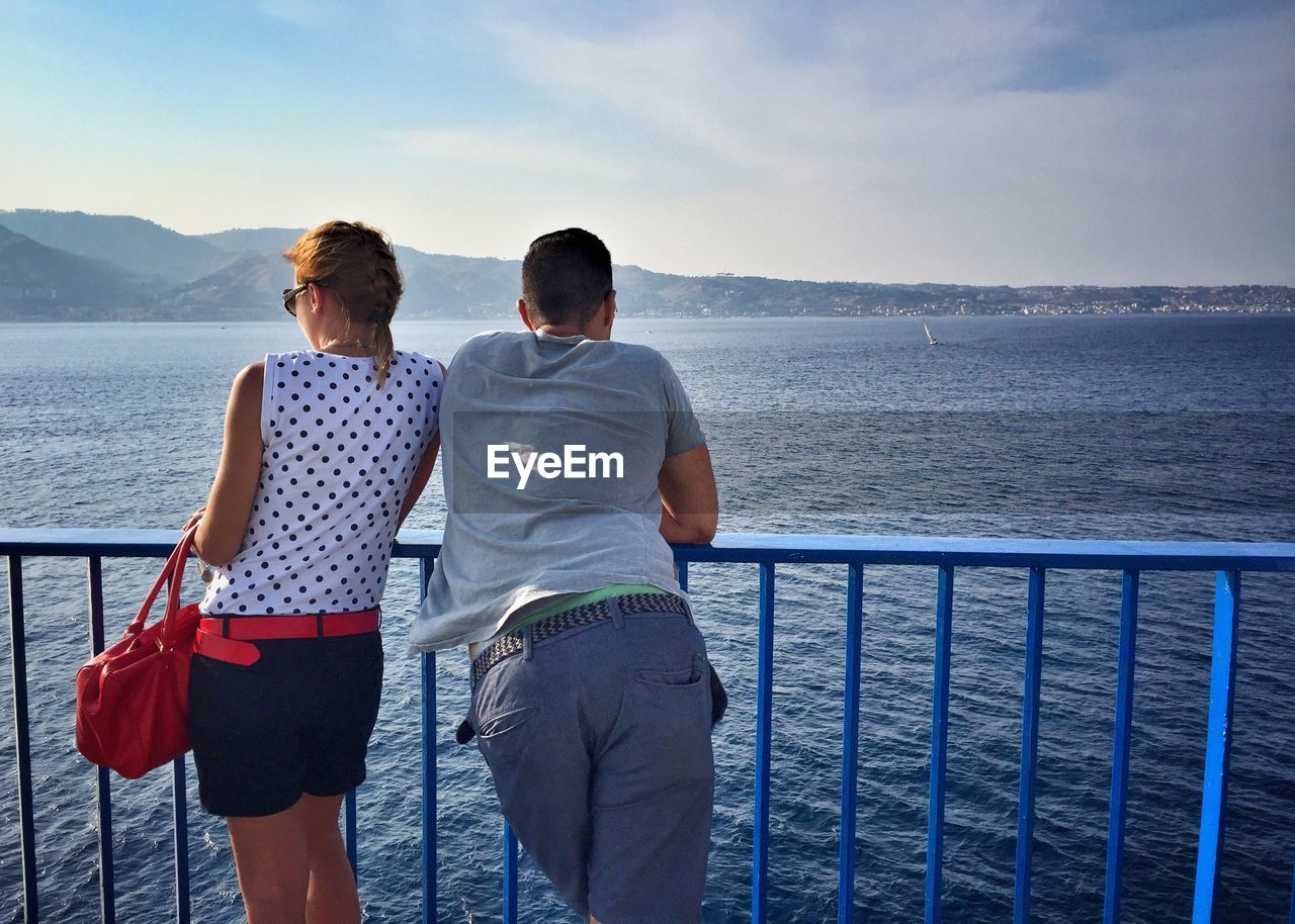 Rear view of couple standing by railing of ferry on sea against sky