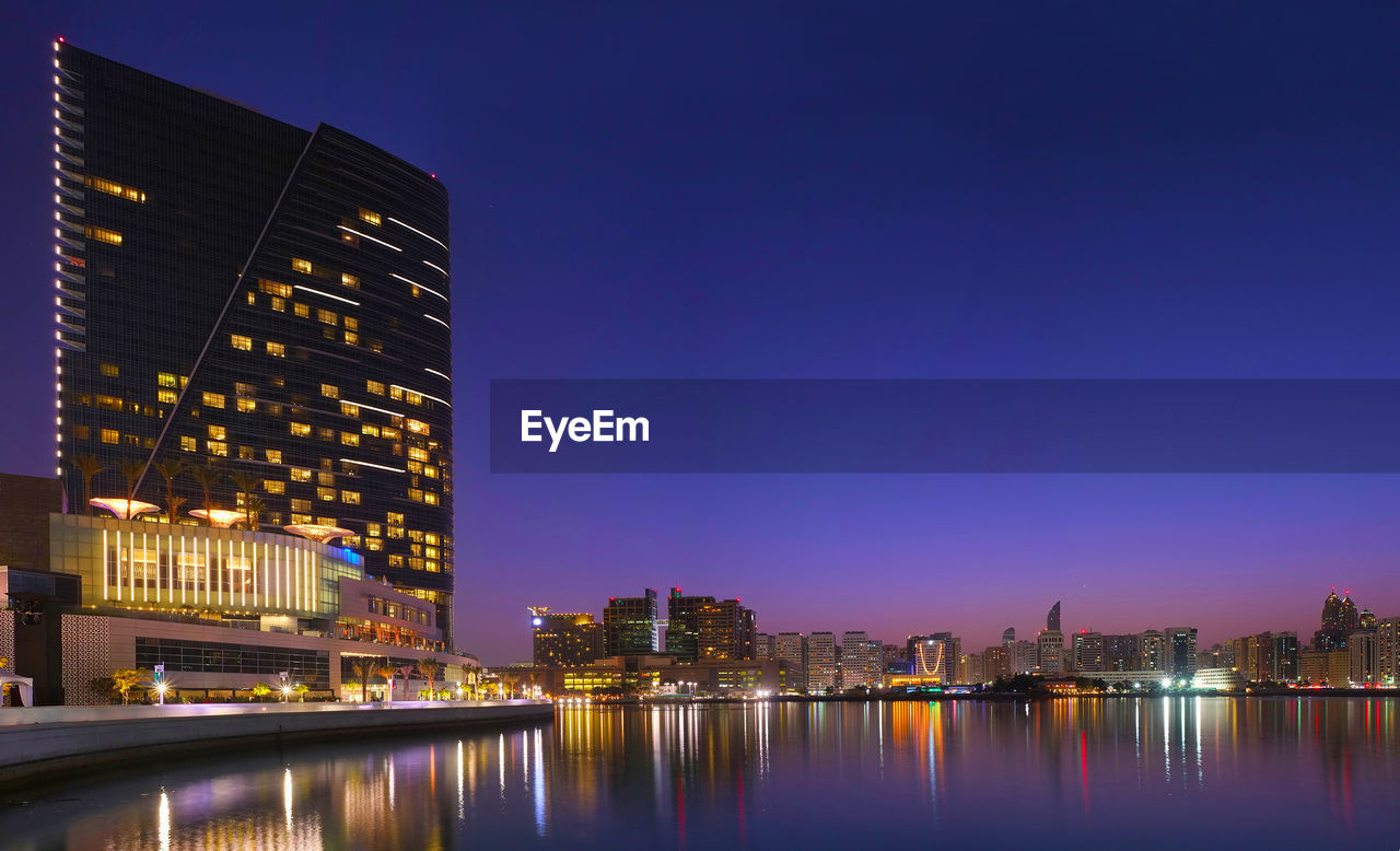 ILLUMINATED MODERN BUILDINGS BY RIVER AGAINST SKY AT NIGHT