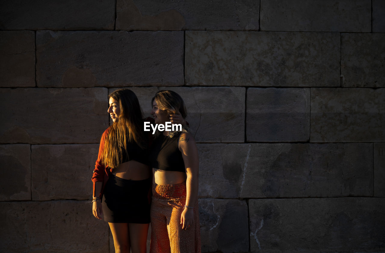 Two girls standing against stone wall during sunset