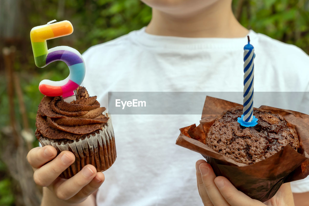 A boy in a white t-shirt holds sweets with candles on the background of the garden.