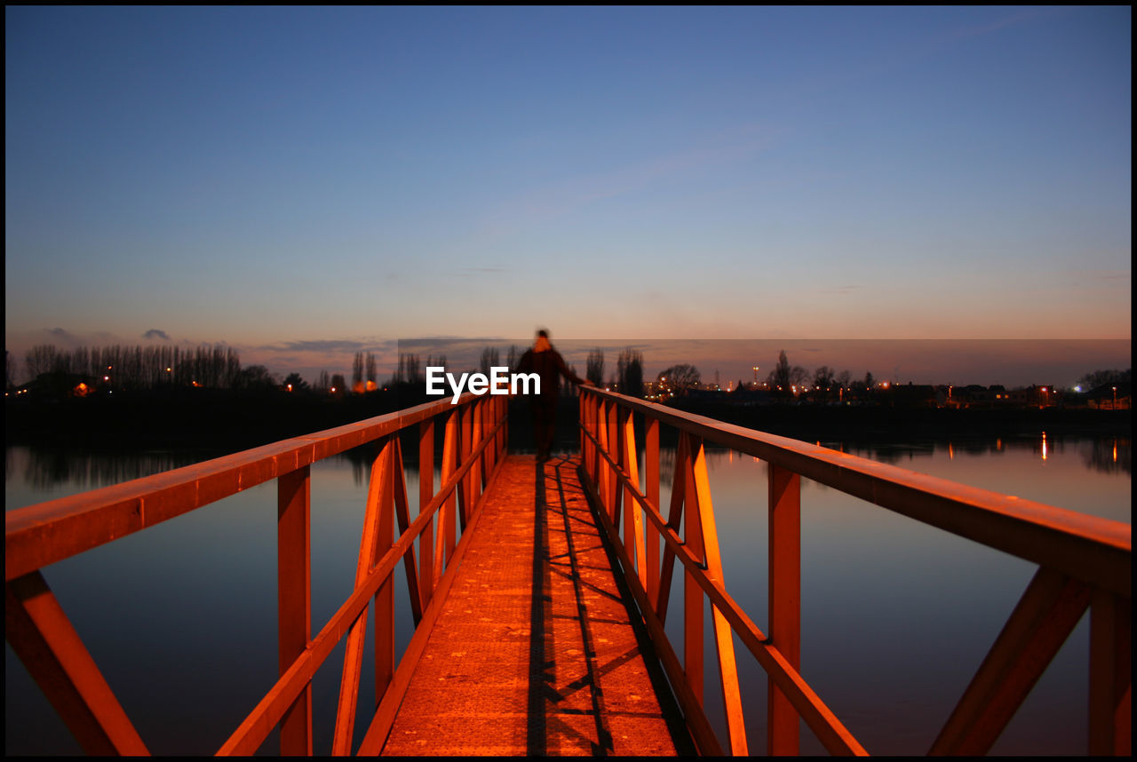 Blurred motion of person walking on footbridge over river against sky at night