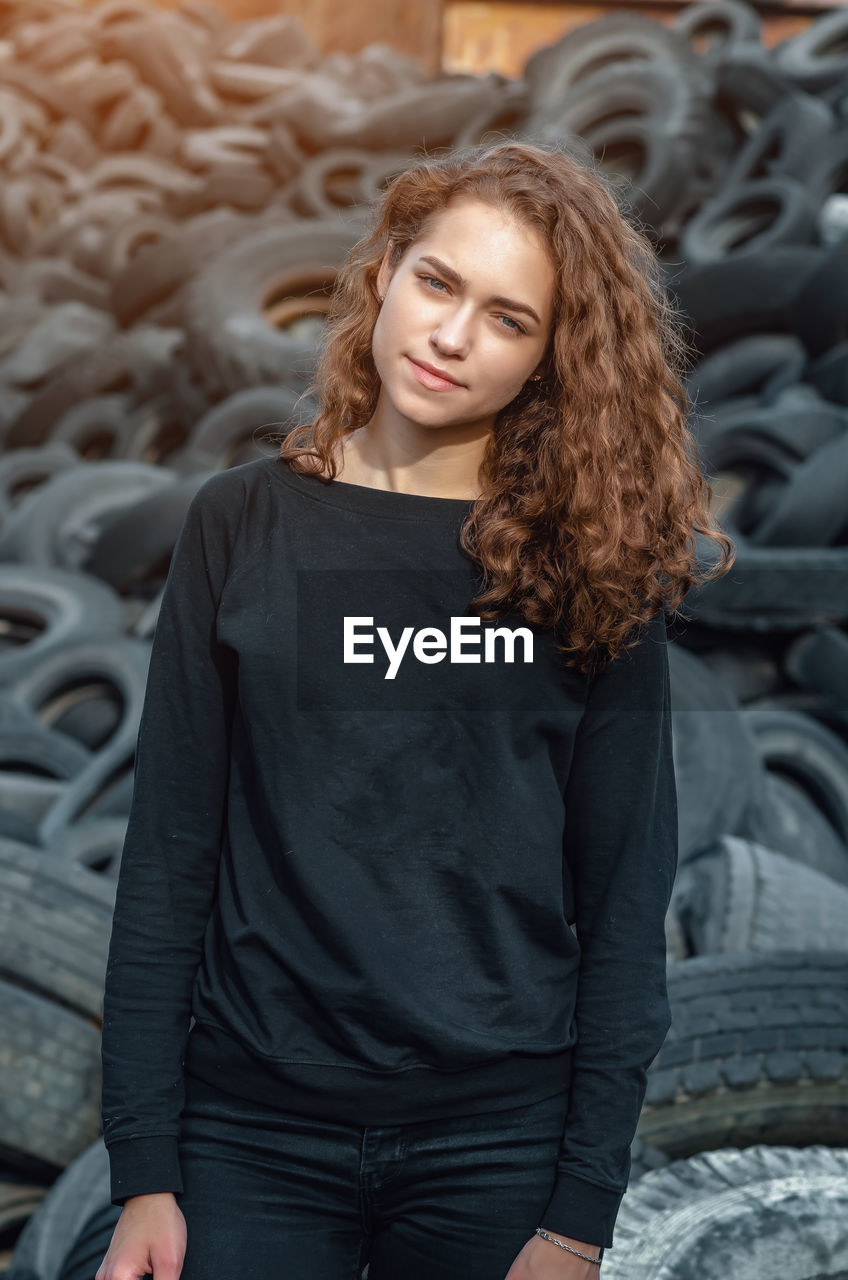 Cheerful curly-haired girl standing against a landfill of used car tires