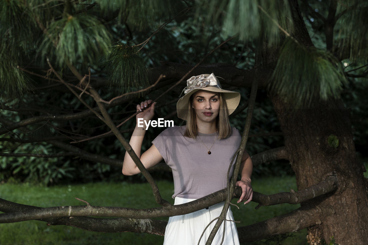 PORTRAIT OF YOUNG WOMAN WEARING HAT STANDING IN FOREST