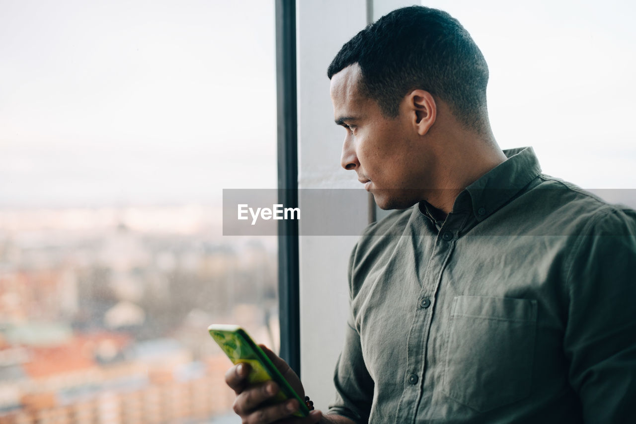 Businessman standing with smart phone while looking through window at creative office