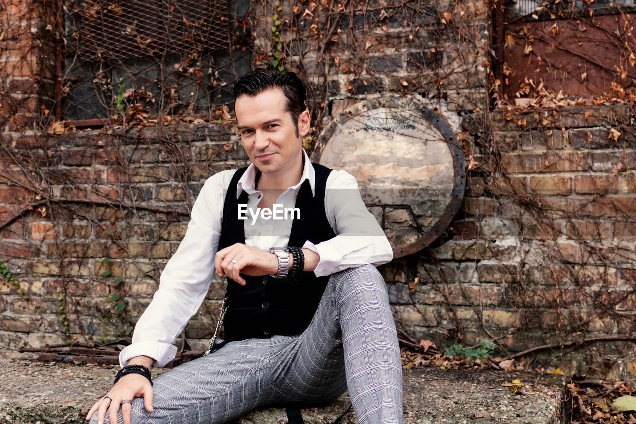 Portrait of happy well dressed man sitting against brick wall and looking at camera.