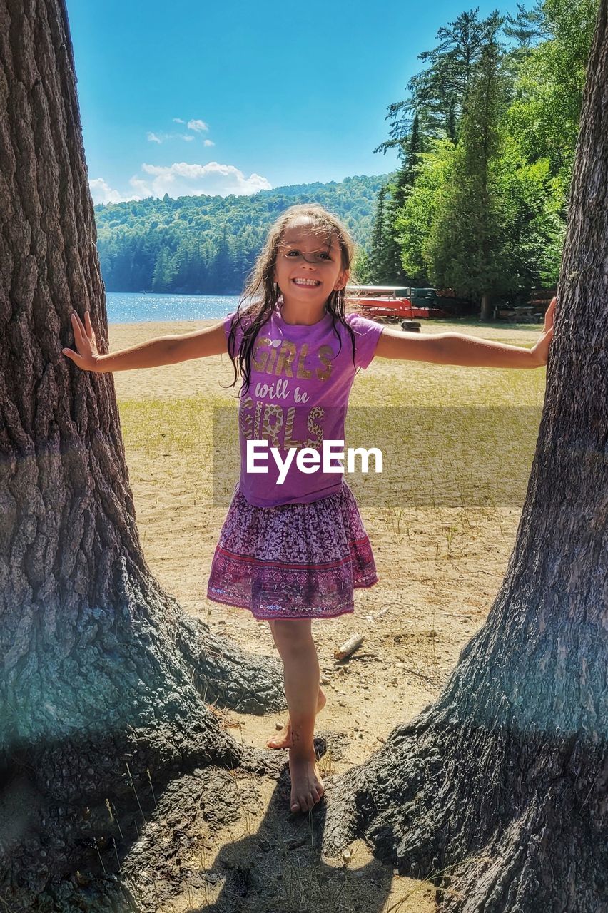 one person, full length, tree, childhood, child, women, dress, nature, plant, standing, female, blue, spring, front view, leisure activity, land, tree trunk, day, trunk, casual clothing, smiling, sunlight, portrait, hairstyle, sky, innocence, long hair, looking at camera, happiness, clothing, person, fashion, outdoors, cute, lifestyles, emotion, footwear, pink, blond hair, shadow, water