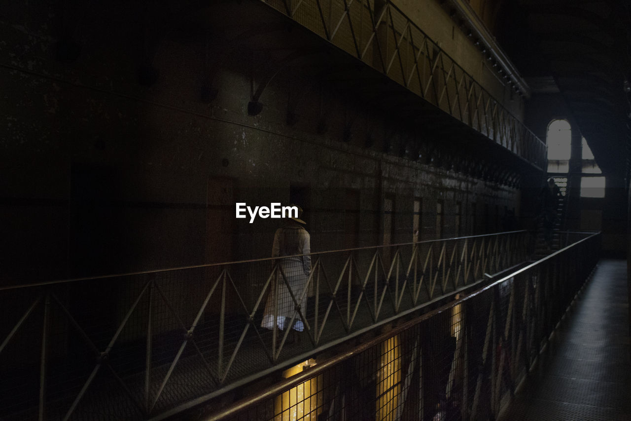 REAR VIEW OF MAN WALKING ON ILLUMINATED BRIDGE IN BUILDING