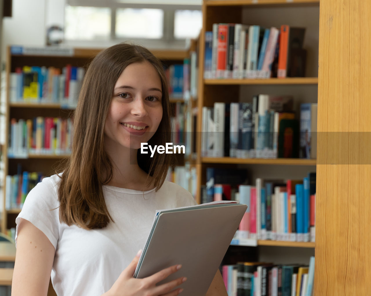 Portrait of smiling teenage girl holding file while standing in library