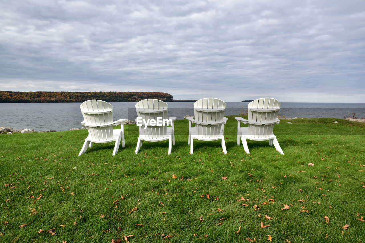 Empty adirondack chairs at grassy lakeshore against cloudy sky