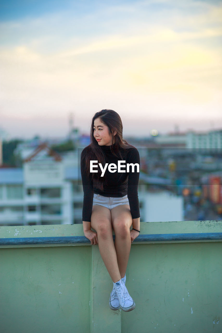 Young woman sitting on railing against cityscape