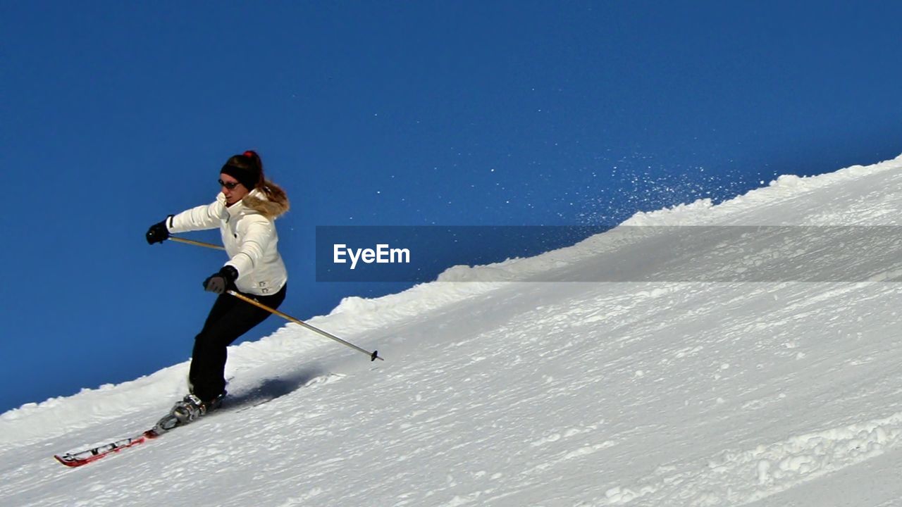 Low angle view of woman skiing on snow covered mountain against clear blue sky