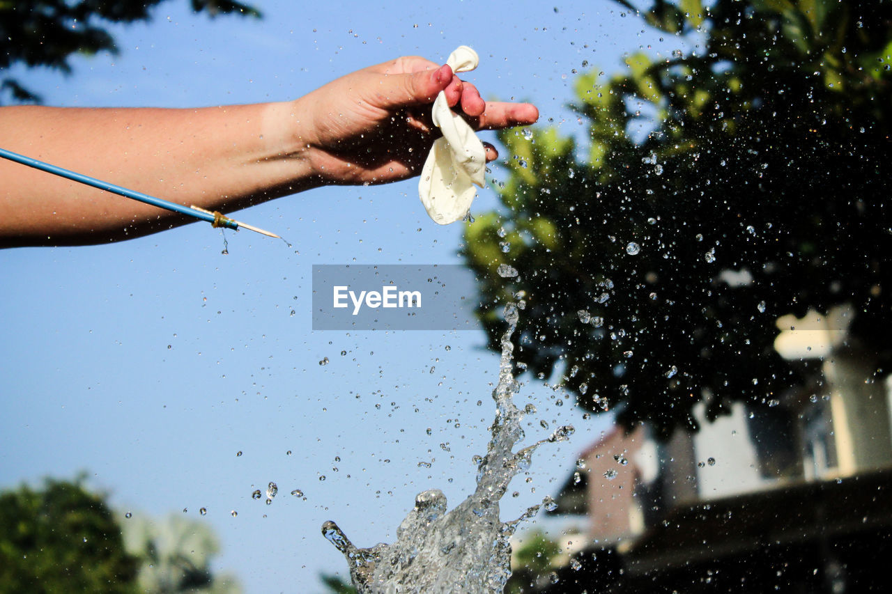 Cropped hand bursting water bomb against clear sky