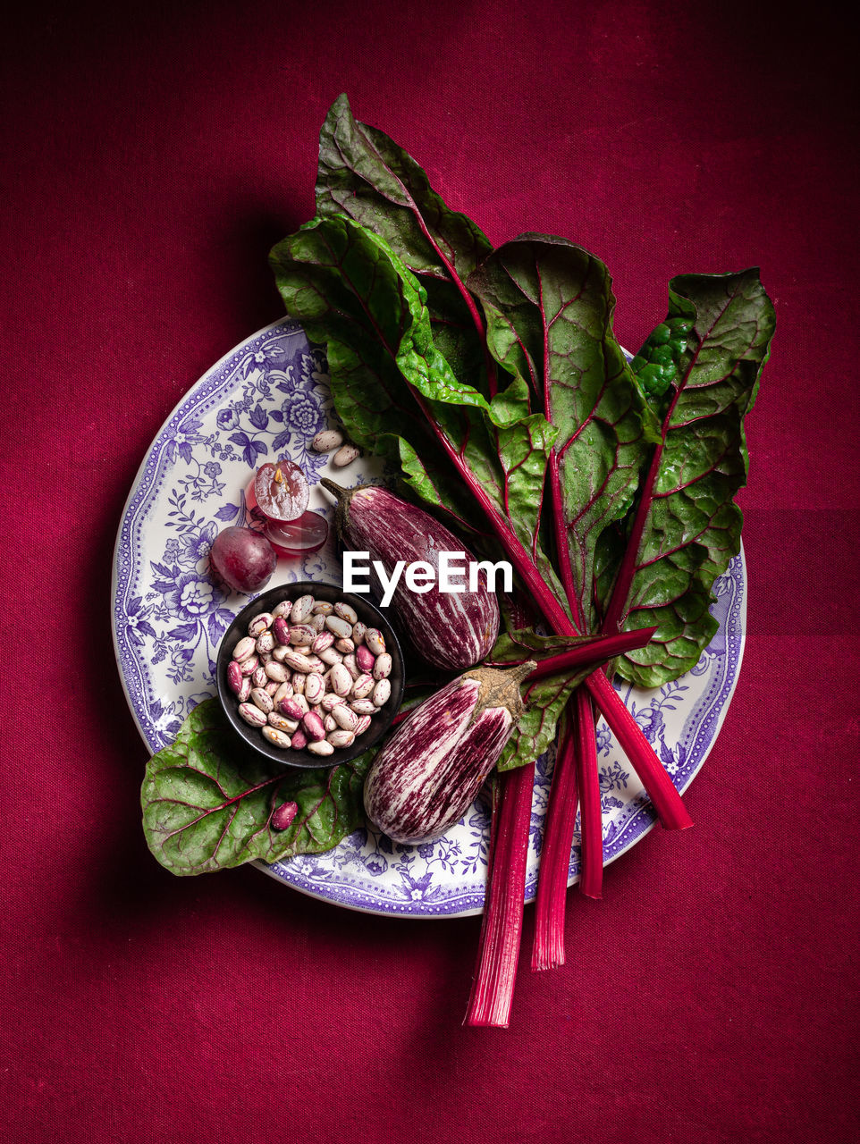 Top view heap of raw chard with purple eggplants served on plate with beans on red background in light studio