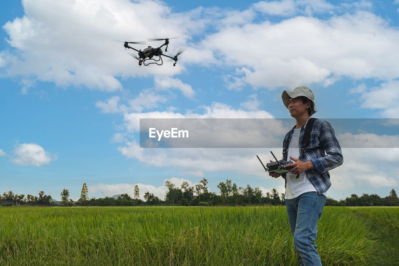 A farmer in a cap stands in a lush wheat field, directing a drone that is flying above the edge.