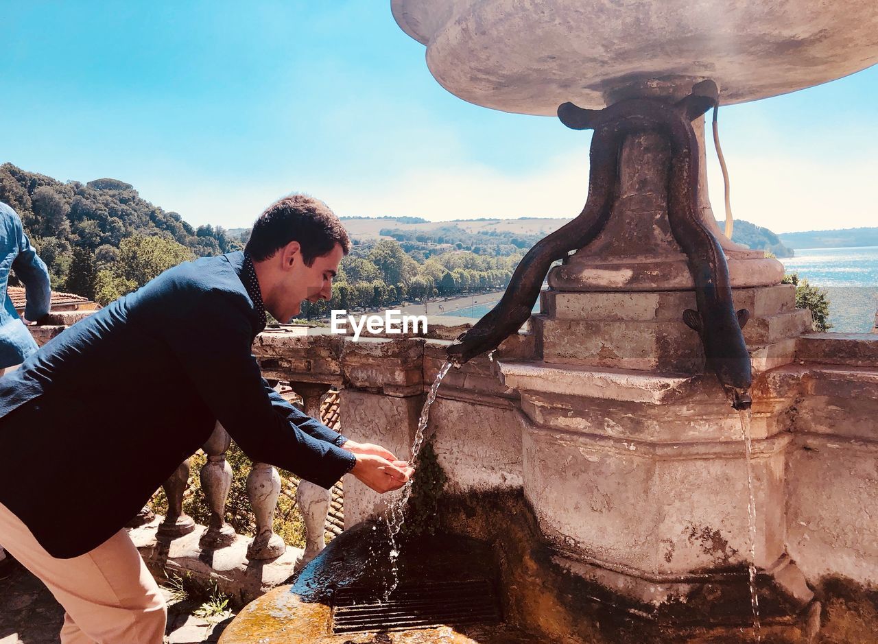 Side view of man gesturing below drinking fountain against sky