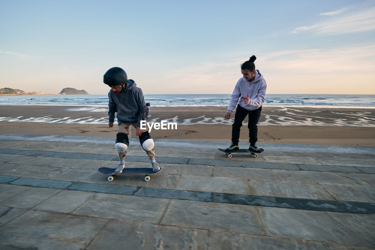 Young active male friends riding skateboards together along promenade on background of sea and sunset sky