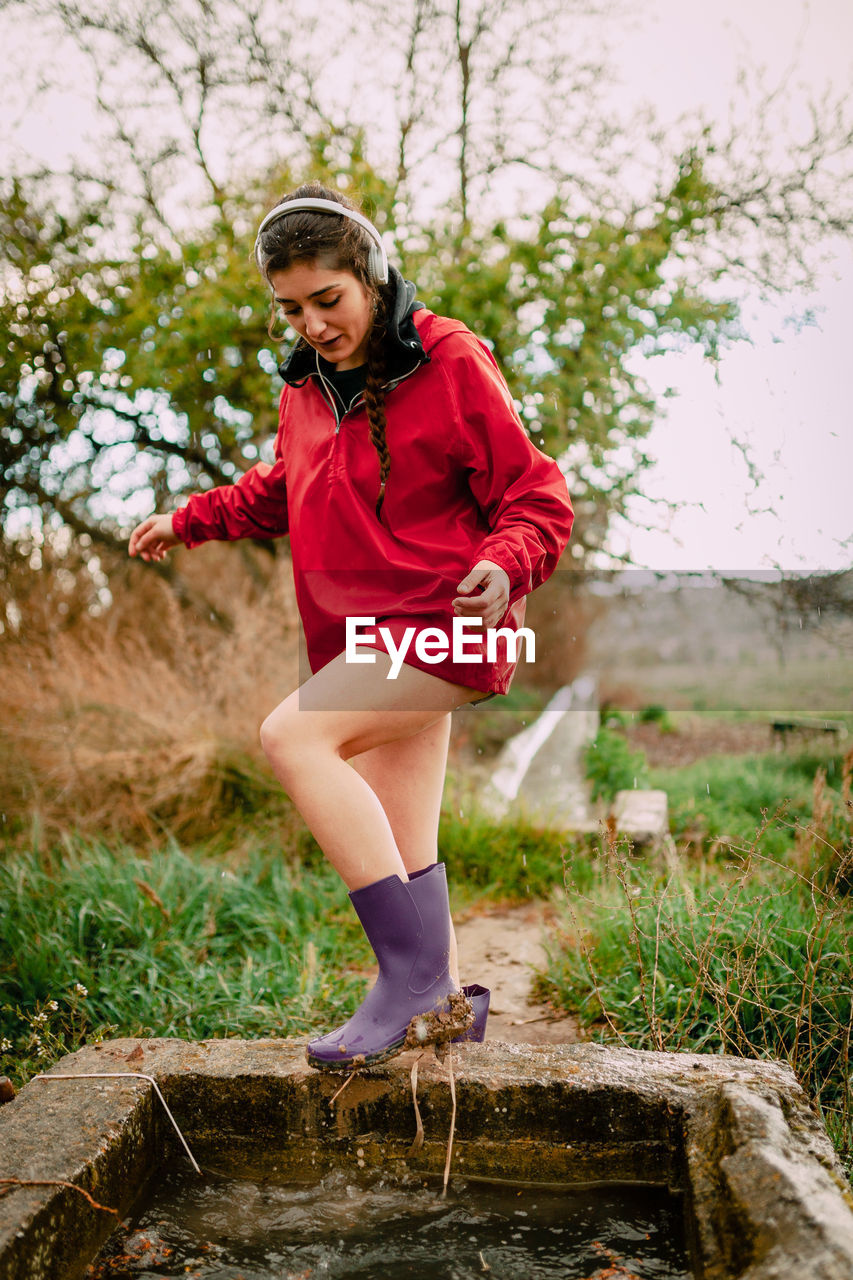 Young woman looking at dirty boot while standing against sky