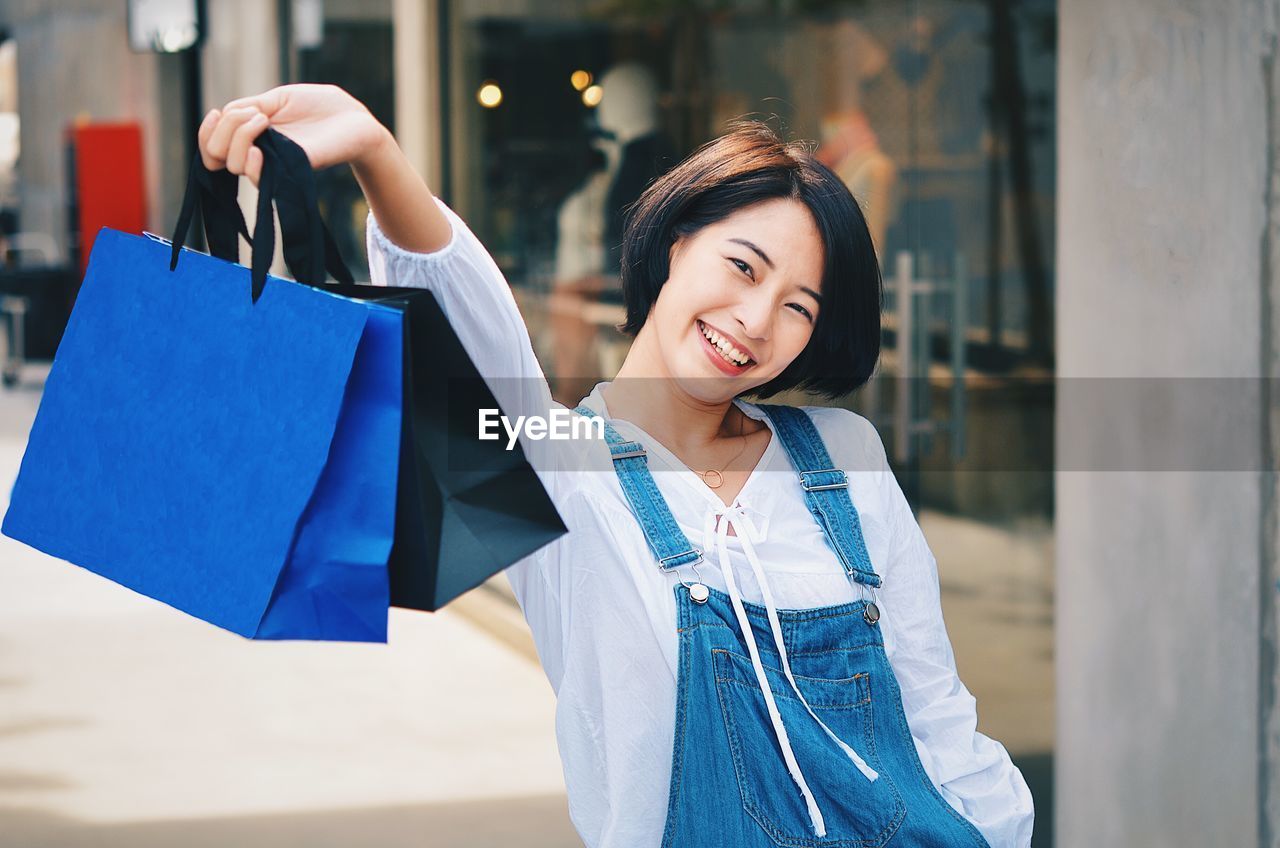 Portrait of happy young woman holding shopping bags while standing at mall