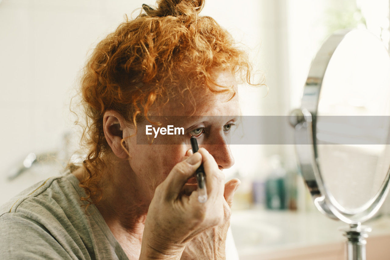 Senior woman looking in mirror and applying eyeliner at home