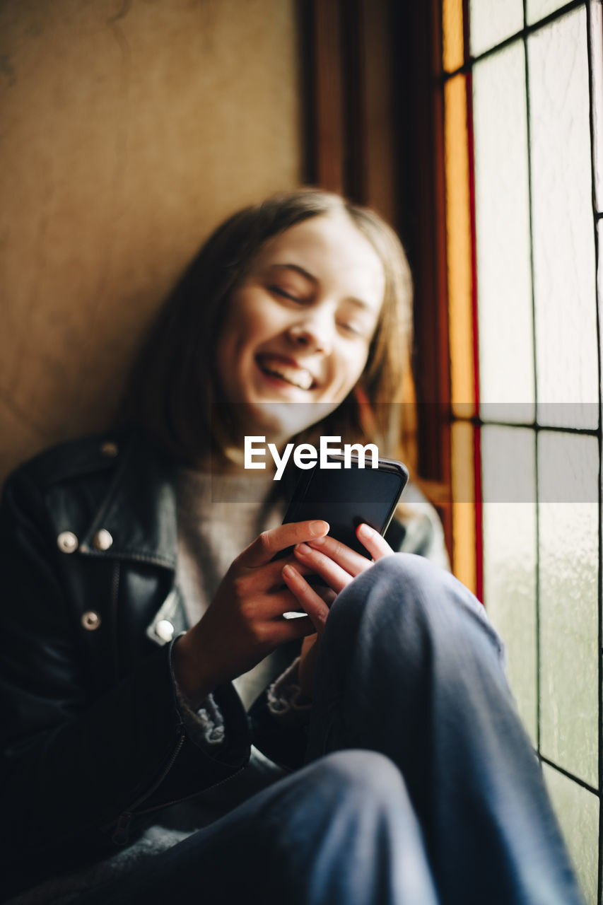 Smiling girl using smart phone while sitting by window at home