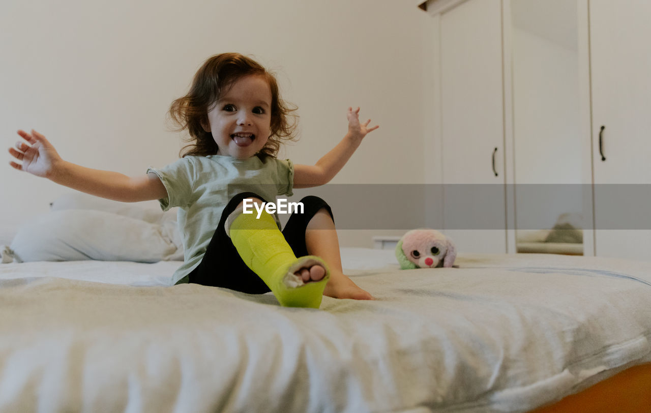 A happy girl with a cast on her leg is sitting on the bed.